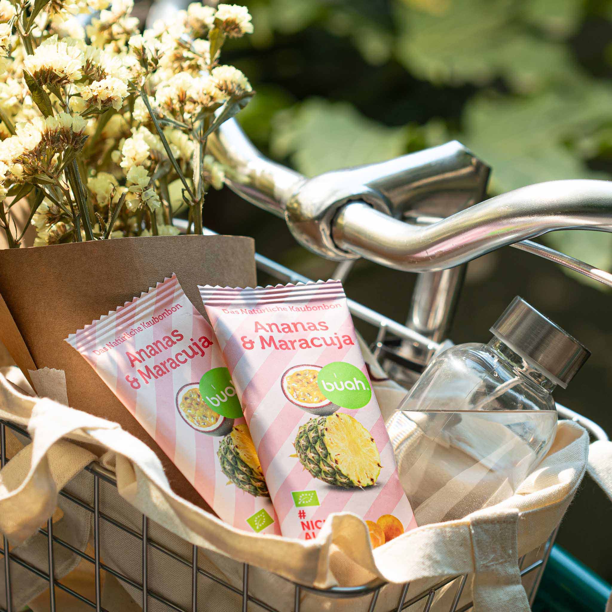 Zwei Packungen buah Ananas-Maracuja Kaubonbon in Fahrradkorb neben frischen Blumen