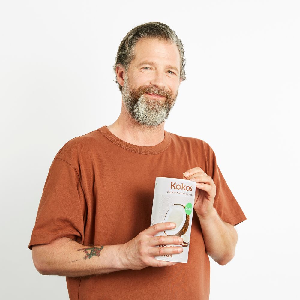 Mann Mitte 40, best Ager, mit braunem Shirt und grau meliertem Bart isst Kokoschips aus einer Dose buah gefriergetrockneter Kokosnuss