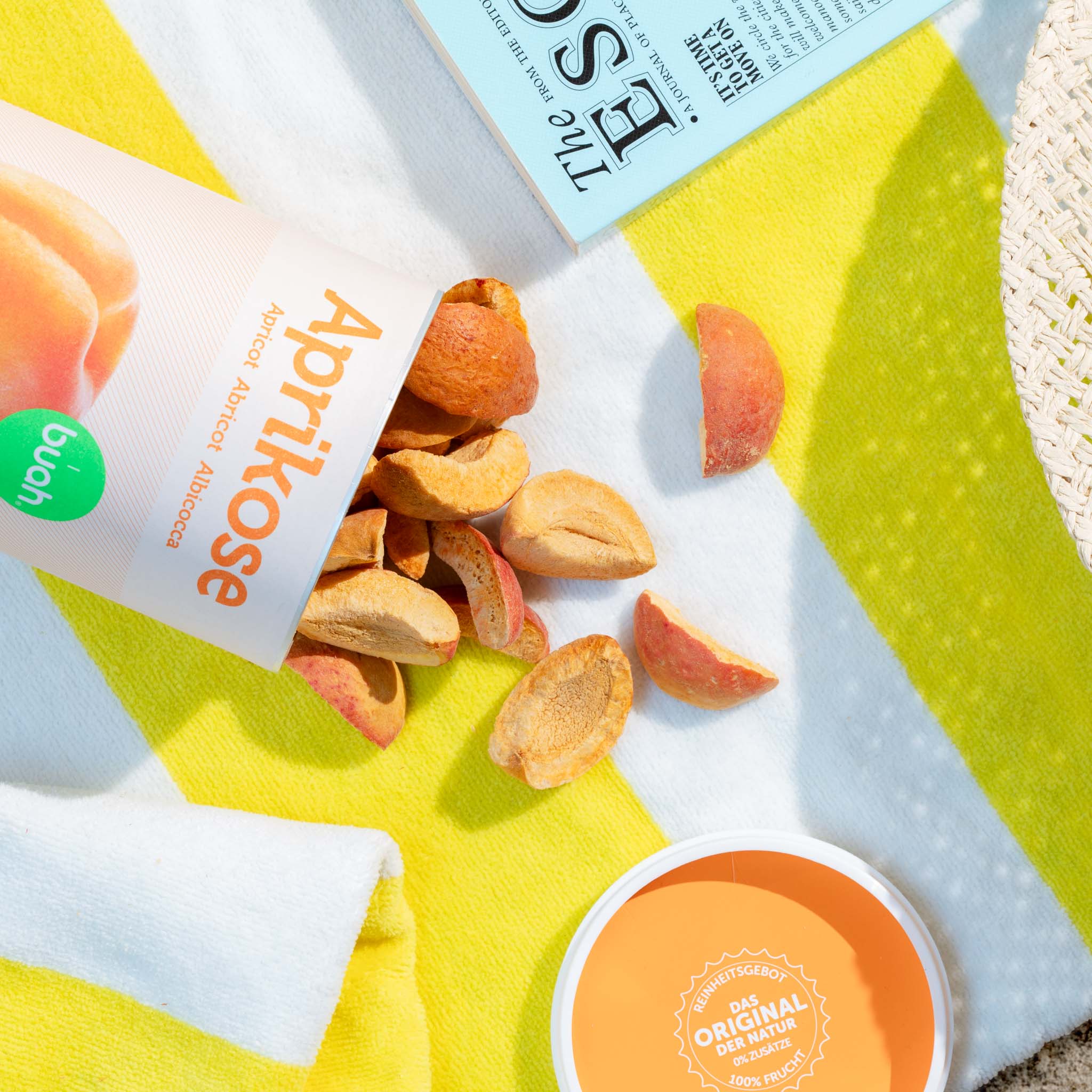 Lattina di albicocca liofilizzata buah capovolta su un telo da mare giallo e bianco, l'albicocca si trova sull'asciugamano accanto al libro