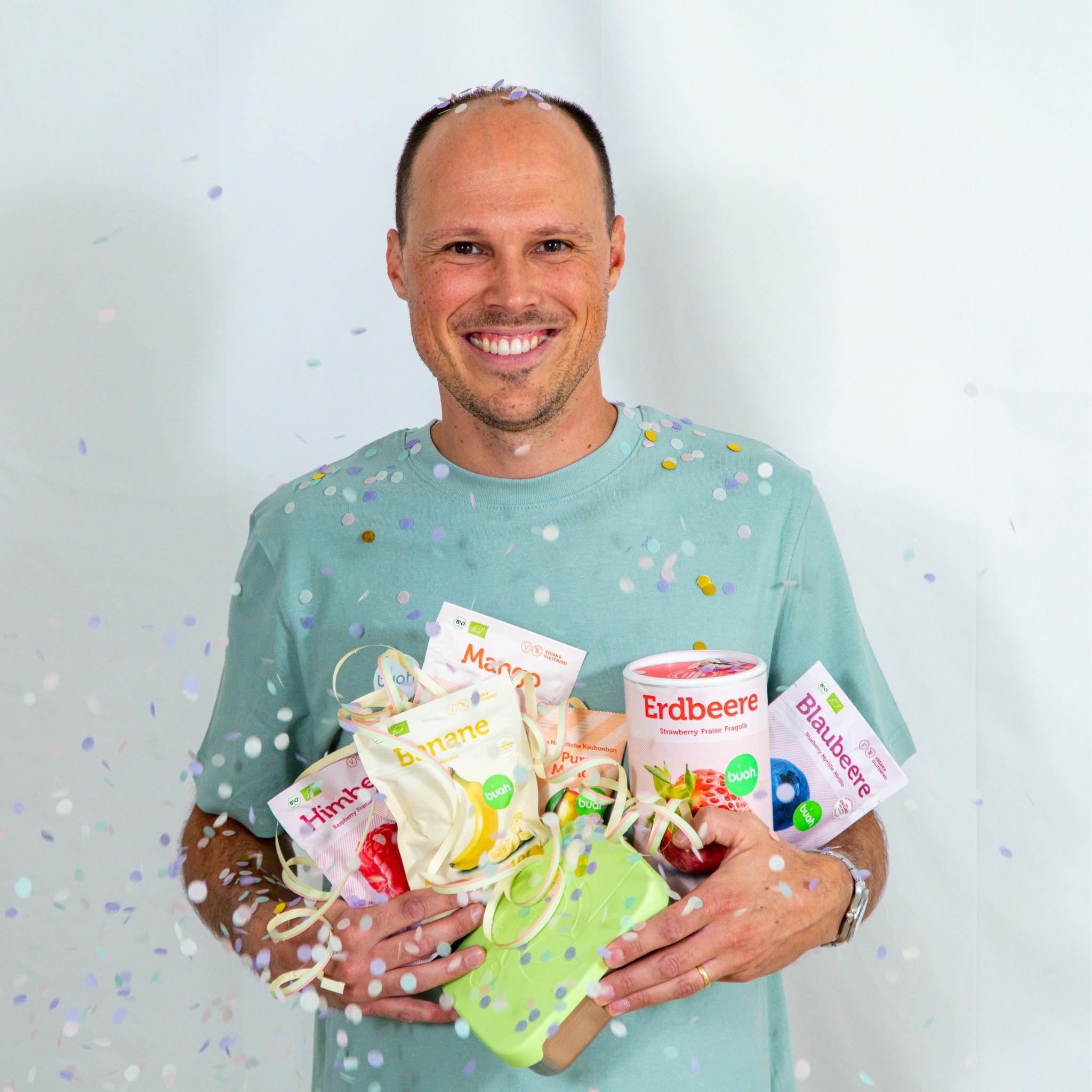 Tobi von buah im gruenem buah Shirt mit buah gefriergetrockneten Fruechten in der Dose im Arm - Konfetti und Luftschlangen im Hintergrund fuer den buah Birthday
