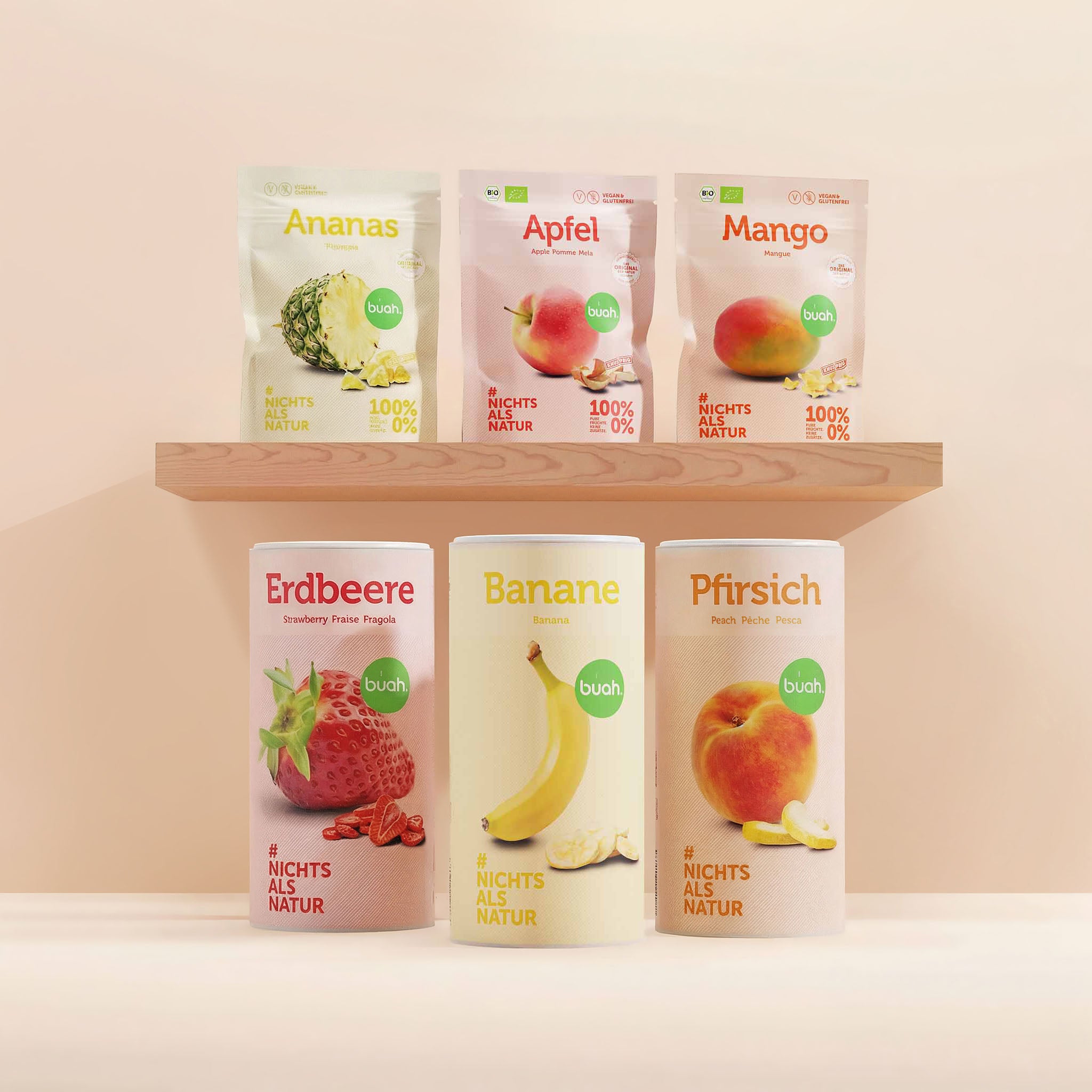 buah Kids-Snack-Set. Ein Regal aus hellem Holz mit drei Snackpacks gefriergetrocknete FrĂĽchte der Sorten Ananas, Apfel und Mango. Darunter stehen auf dem Boden drei groĂźe Dosen der Sorten Erdbeere, Banane und Pfirsich vor einem beigefarbenen Hintergrund.