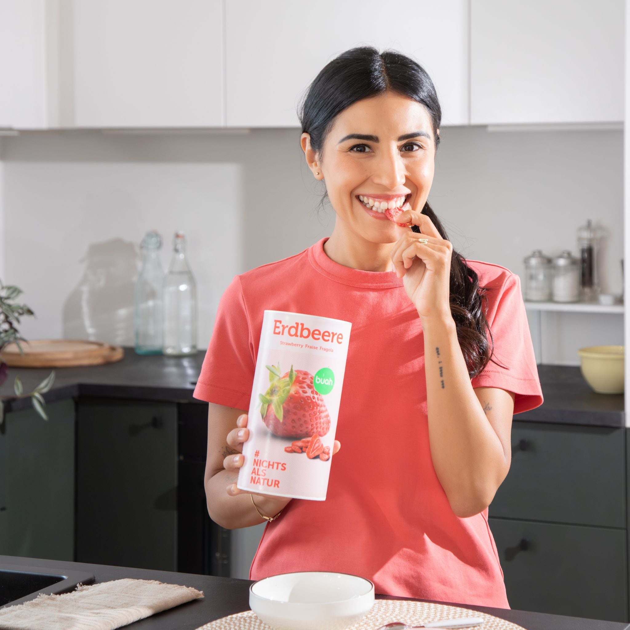 Junge Frau mit langen dunklen Haaren snackt ein Stueck gefriergetrocknete Erdbeere aus einer Dose buah Erdbeere und lacht in die Kamera