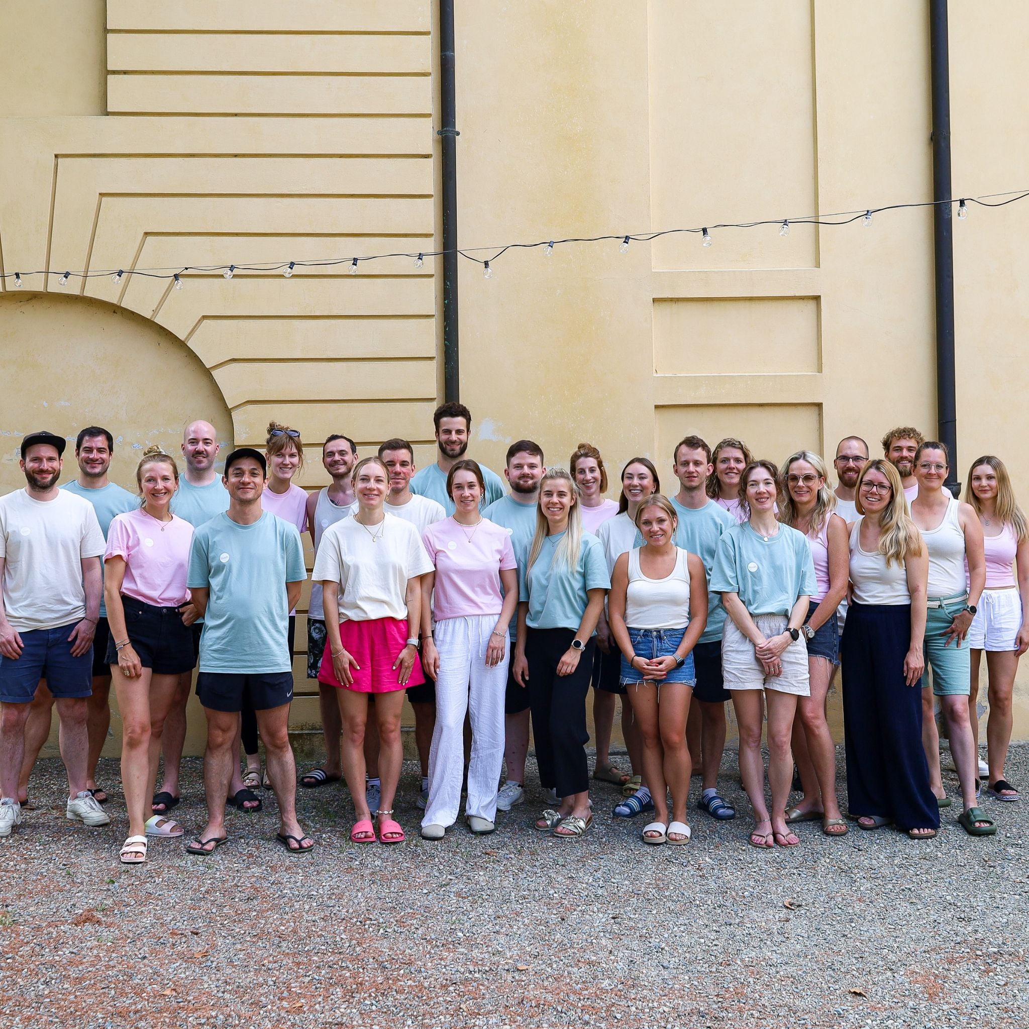 Bild des buah Teams vor einer gelben Wand, viele Frauen und Maenner in Pastell Farben Shirts, im Sommer