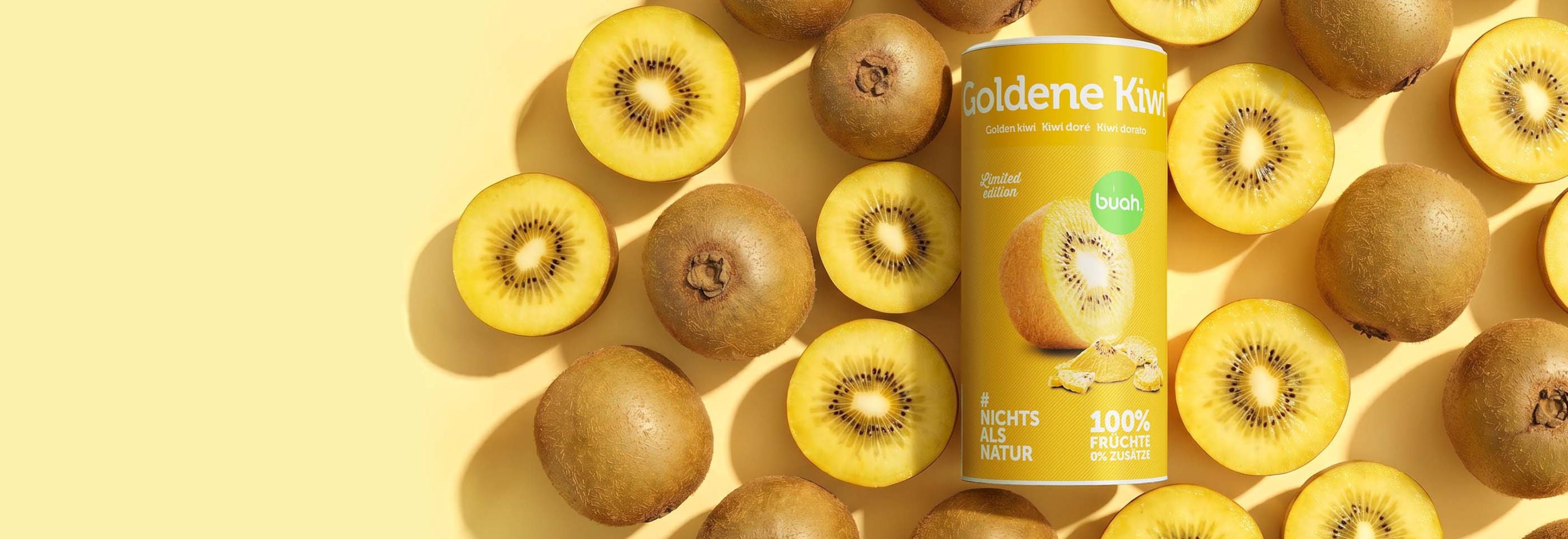 Eine flach von oben fotografierte Anordnung im Querformat auf hellgelbem Hintergrund. Im Zentrum steht eine gelbe Dose buah gefriergetrocknete Goldene Kiwi (Limited Edition), umgeben von zahlreichen frischen, ganzen und halbierten Goldenen Kiwis.