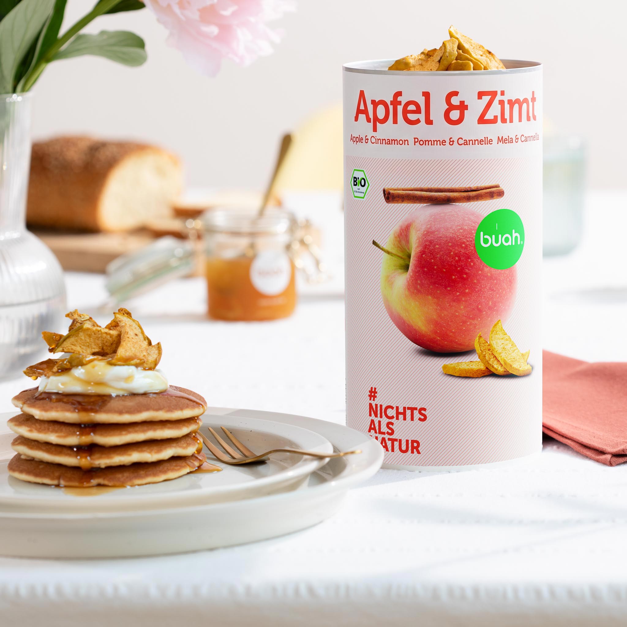 Ein gedeckter Frühstückstisch im hellen Sonnenlicht. Eine große Dose buah gefriergetrocknete Früchte der Sorte Apfel & Zimt steht neben einem Teller mit gestapelten Pancakes, die mit Joghurt, Sirup und knusprigen Apfelstücken garniert sind.