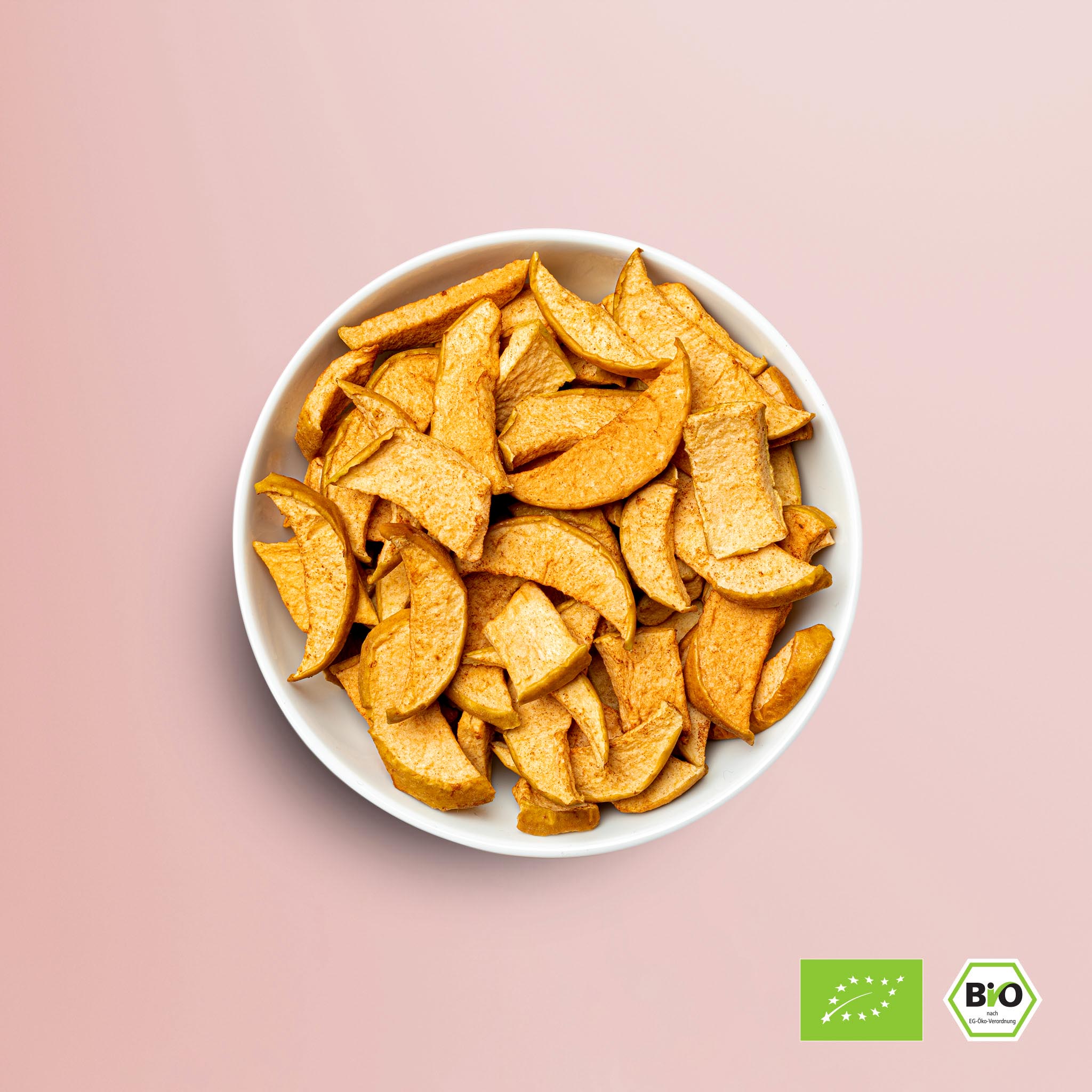Weiße Bowl mit buah gefriergetrocknetem ApfelZimt in gelblicher Farbe mit braunem Staub, vor rosa Hintergrund, BIO Logo in der Ecke der Abbildung