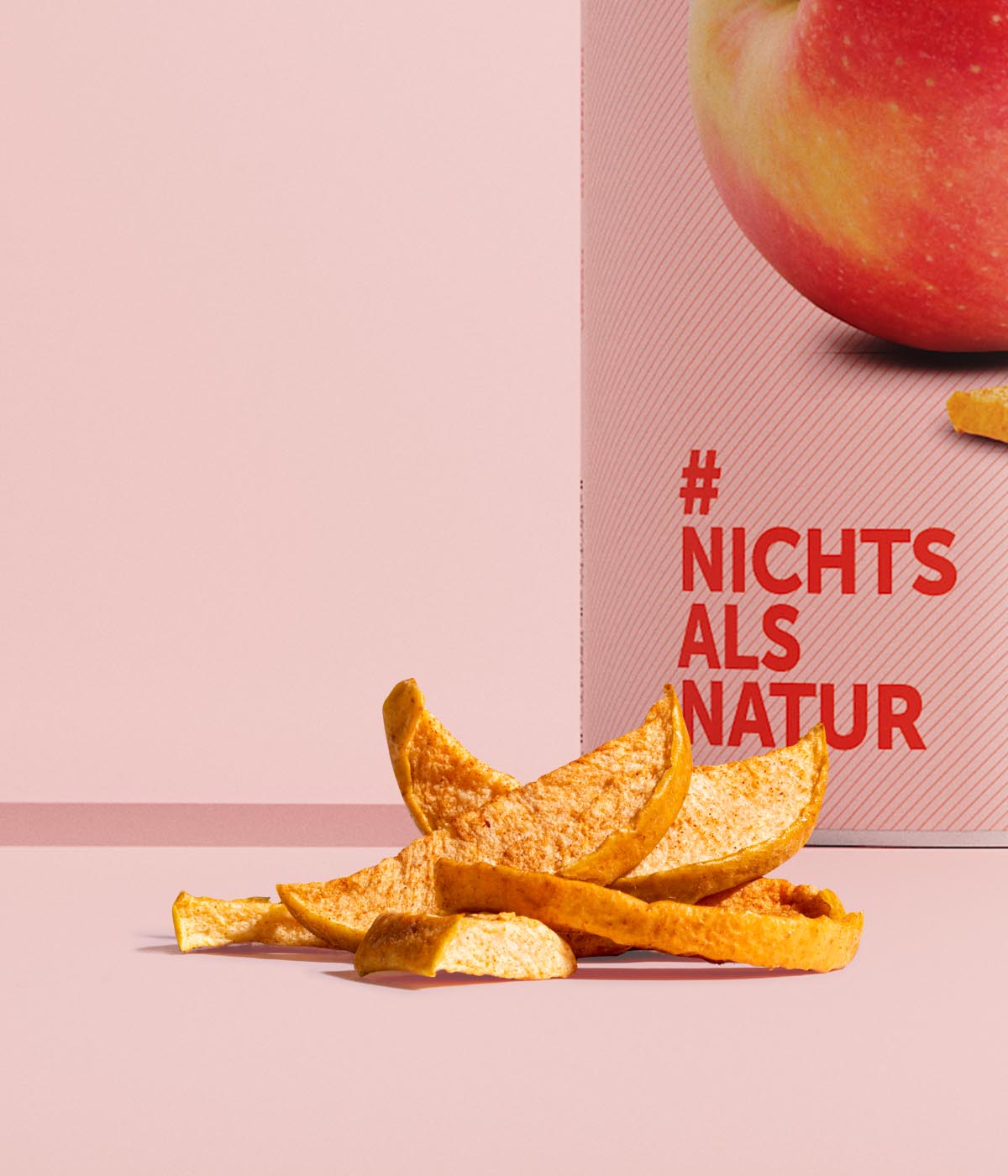 Ausschnitt und Nahaufnahme einer Dose buah ApfelZimt, wenige Stuecke gefriergetrockneter ApfelZimt vor der Dose vor rosa Hintergrund