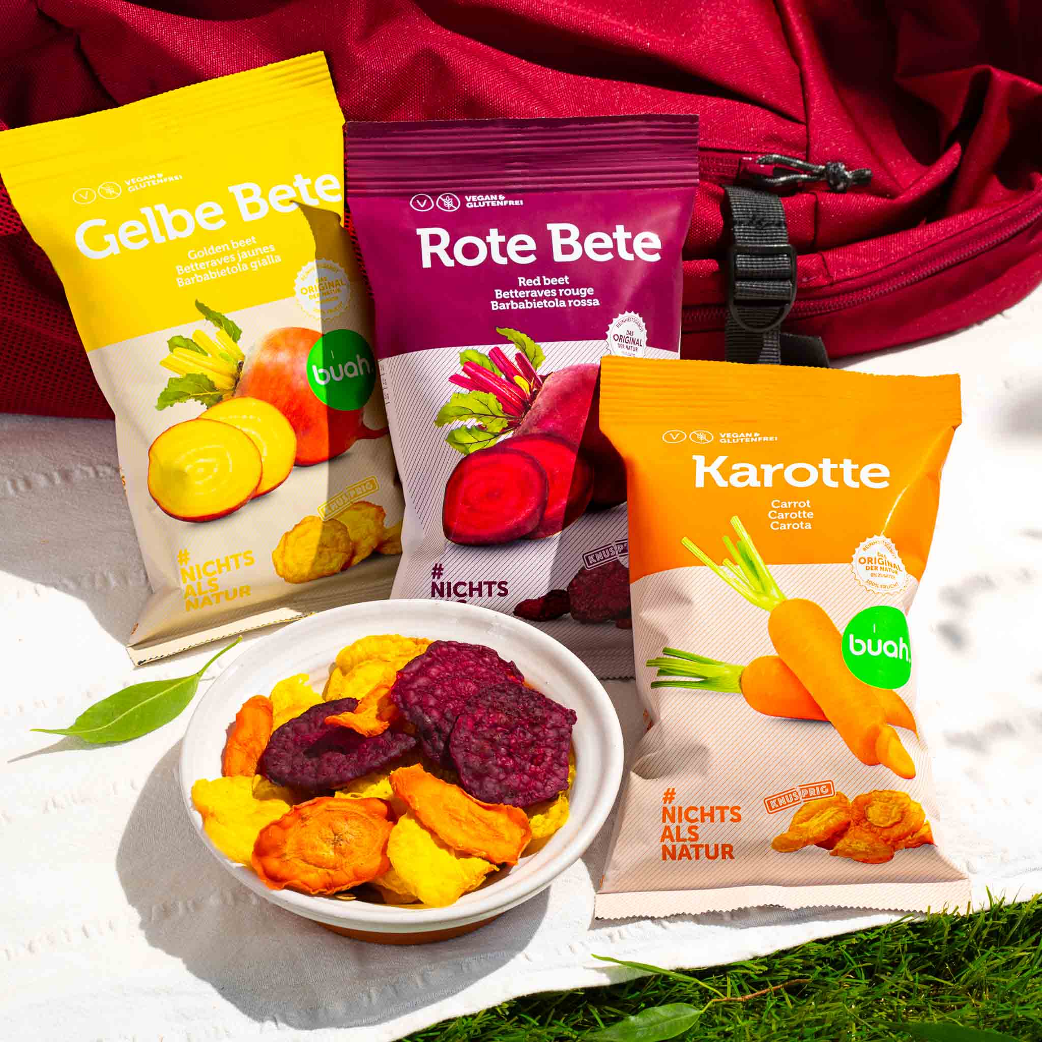 buah Gemuesechips in den Sorten Rote Bete, Gelbe Bete und Karotte liegen neben einem Wanderrucksack