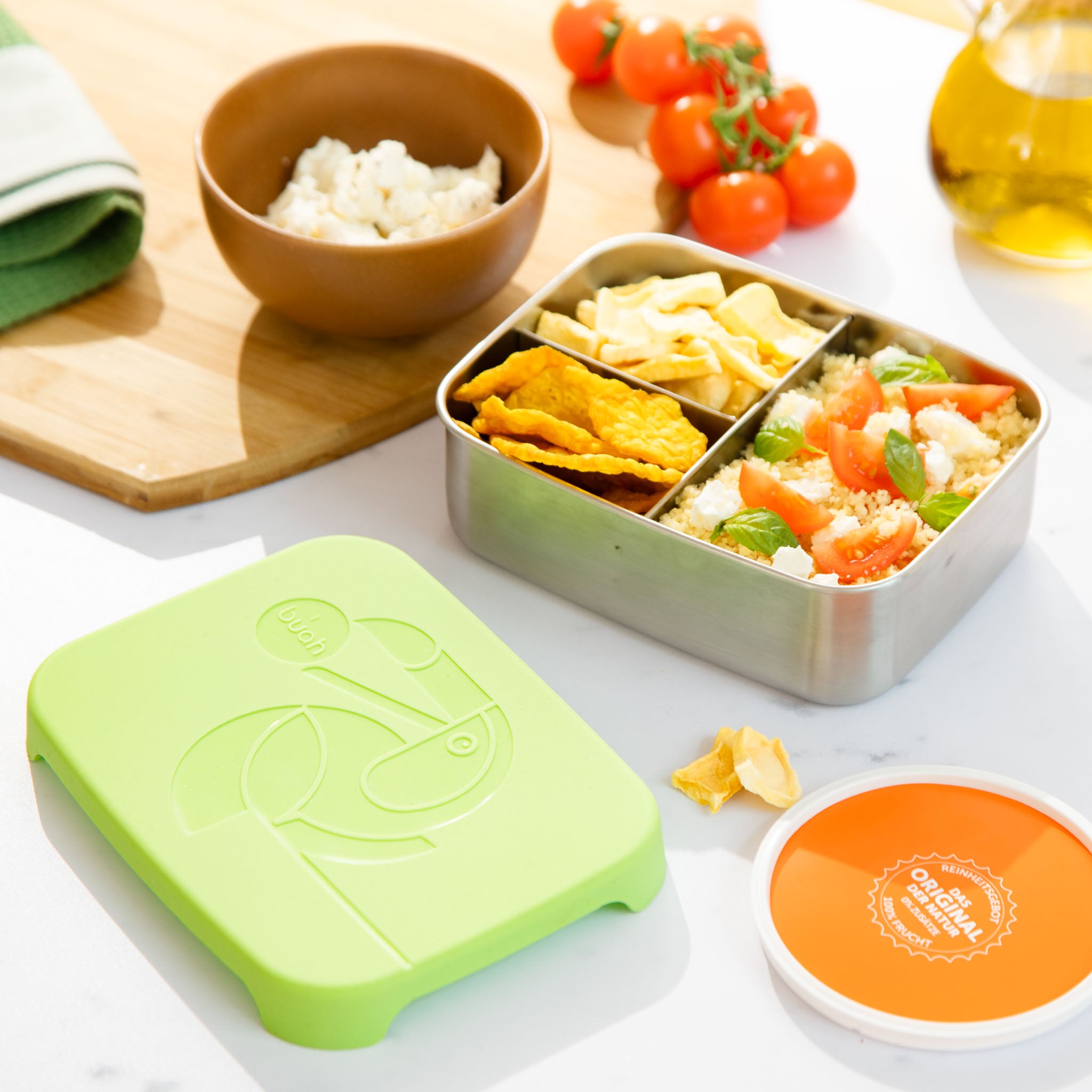 buah Brotdose aus Edelstahl liegt offen auf Esstisch und ist gefuellt mit buah Gemuesechips und buah Mango, sowie Couscous Salat. 