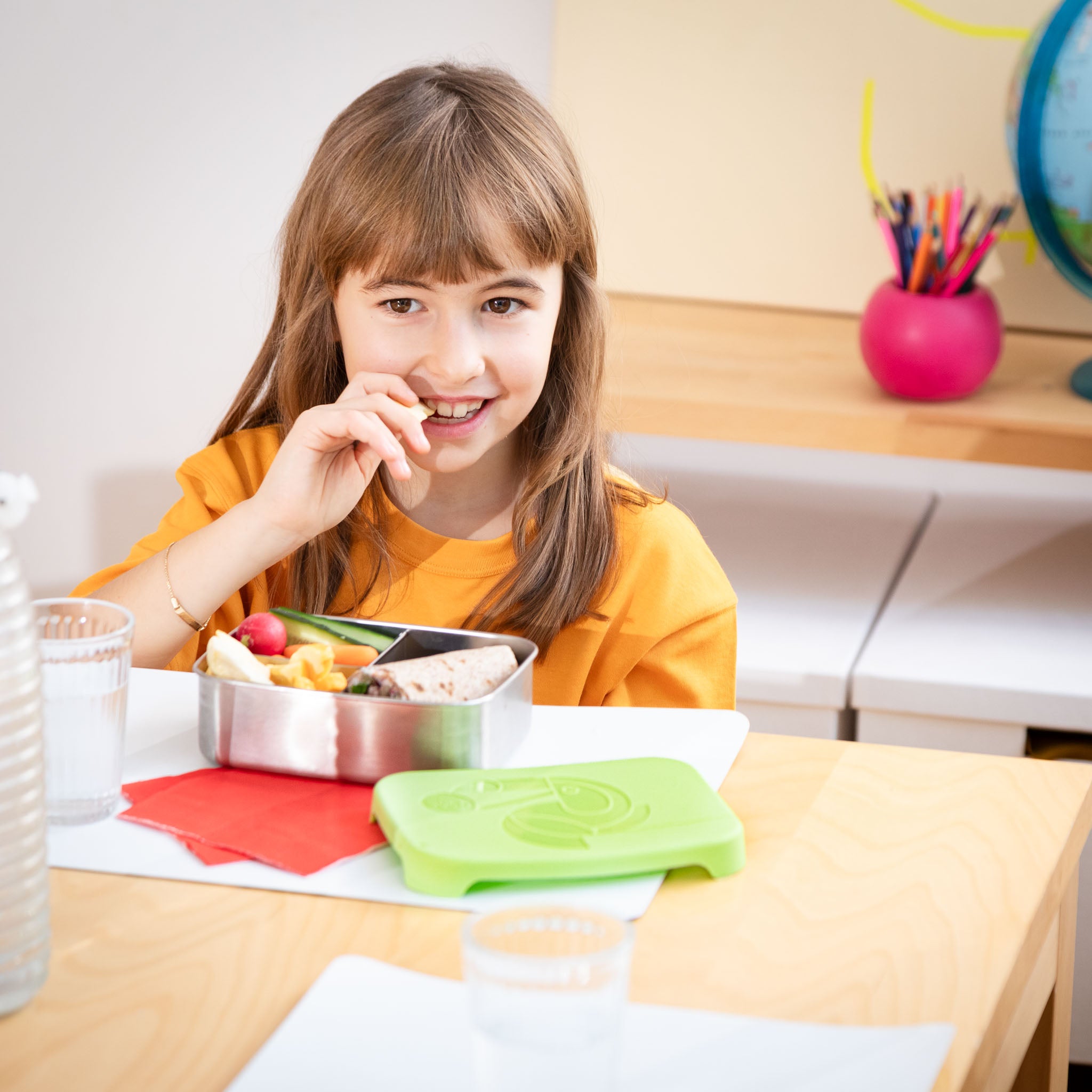 Kind in der Schule sitzt am Tisch und isst Brotzeit aus der buah Lunchbox und lacht in die Kamera. 