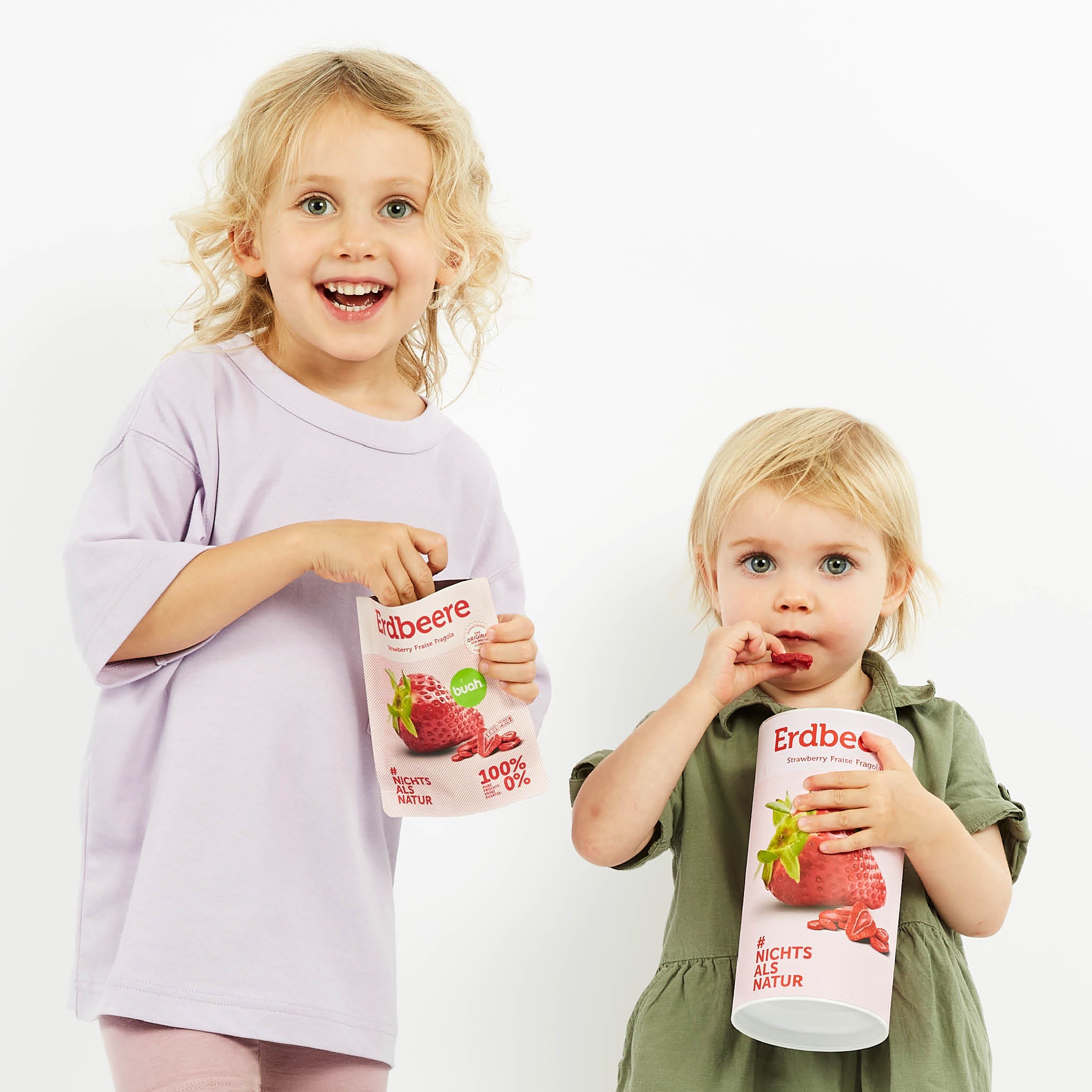 Zwei kleine blonde Kinder mit buah gefriergetrockneter Erdbeere in der Hand lachen in die Kamera. 