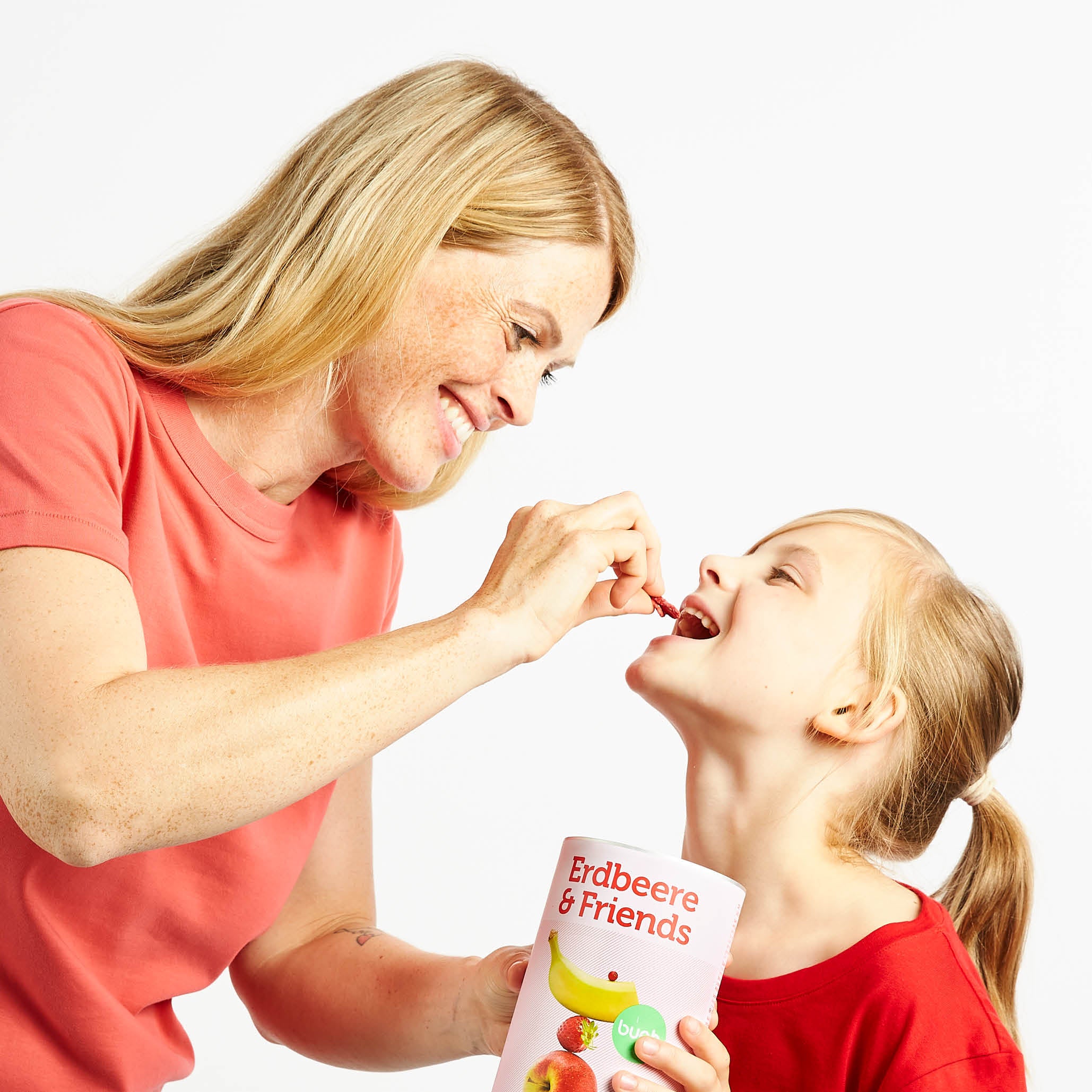Lachende Mama gibt kleiner Tochter Fruechte von buah aus der Dose Erdbeere & Friends