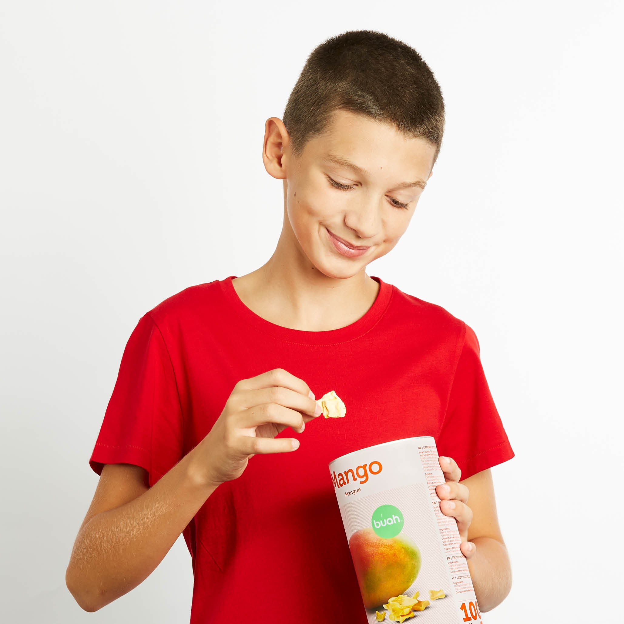 Junge im roten T-shirt isst aus einer Dose buah gefriergetrocknete Mango und lacht
