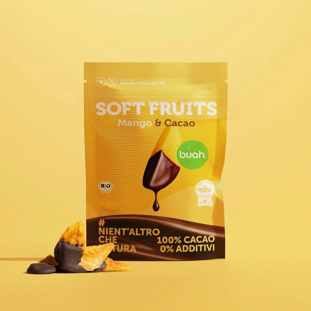 Un sacchetto giallo di buah Soft Fruits al gusto Mango & Cacao si trova su uno sfondo giallo. Davanti ci sono alcuni morbidi pezzi di mango ricoperti di cioccolato.