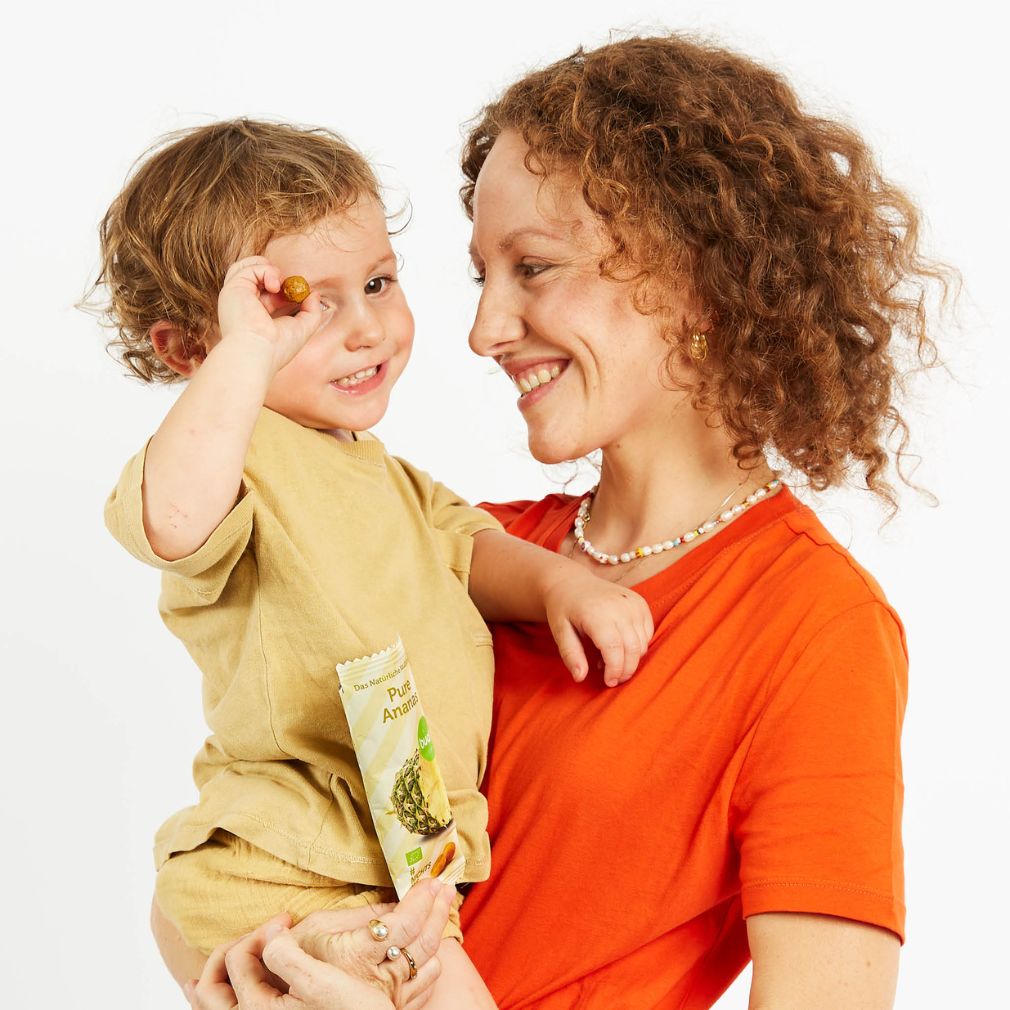 Junge Frau mit kurzen braunen Locken und rotem Shirt hat Kleinkind auf dem Arm, dass buah Frucht Kaubonbons isst und dabei lacht