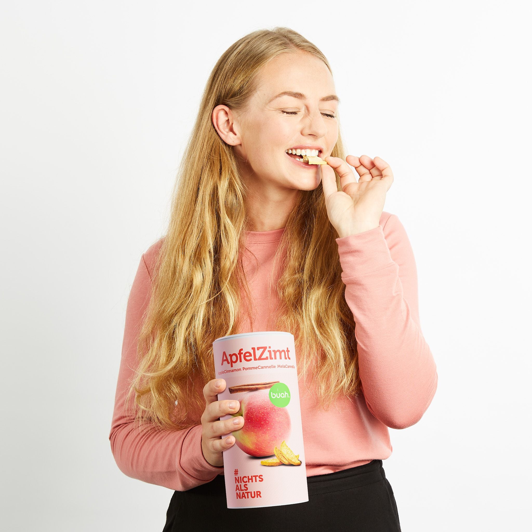 Junge Frau mit langen blonden Haaren und rosa Shirt lacht in die Kamera, mit geschlossenen Augen und snackt buah gefriergetrockneter ApfelZimt