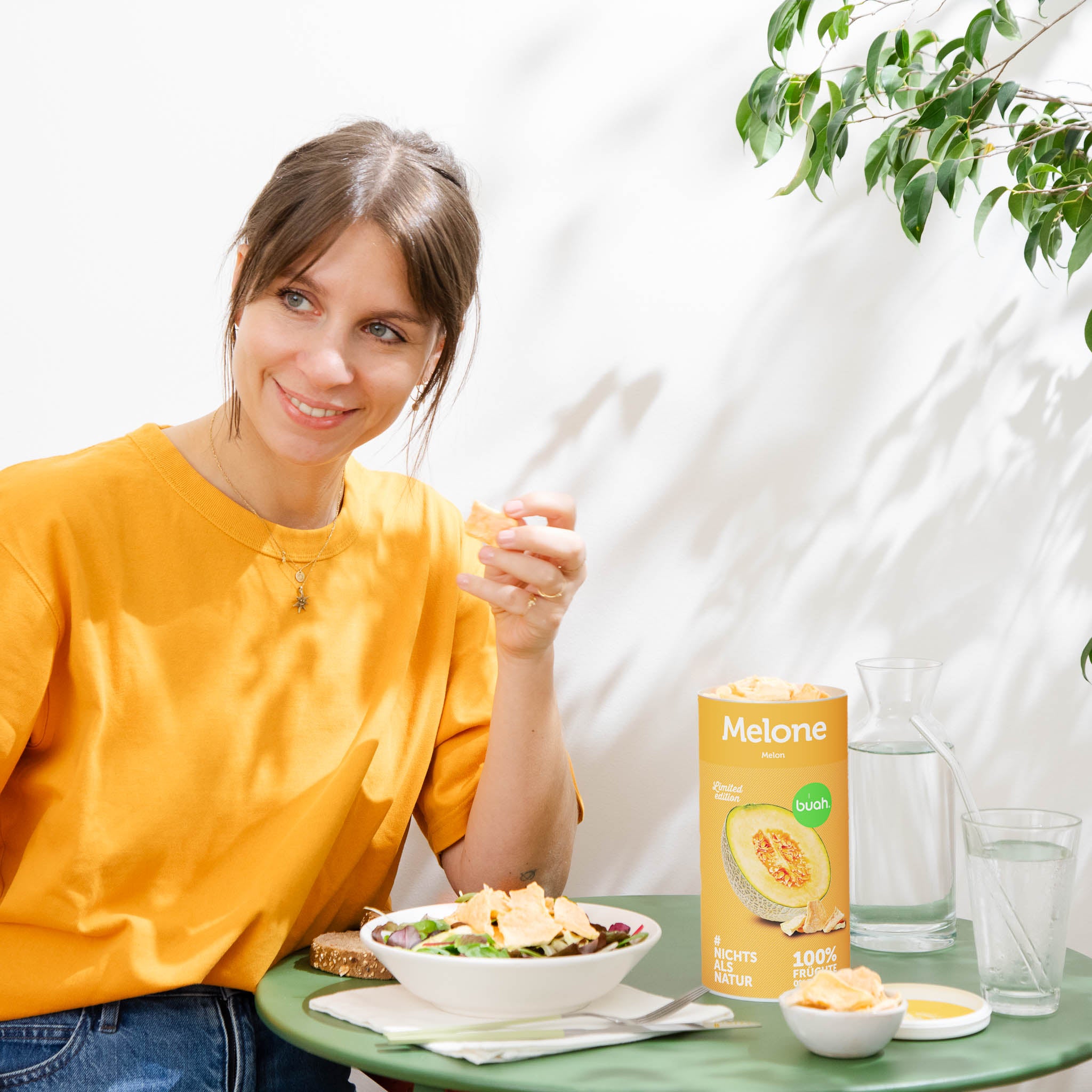 Junge Frau mit orangenem Shirt sitzt an kleinem Tisch und geniesst buah gefriergetrocknete Melone aus der Dose und gibt Melone auf Ihren Salat. 