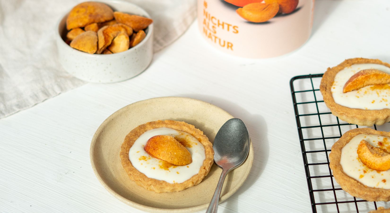 buah Tartletts mit einer Creme und buah Aprikose als Topping auf einem Teller, daneben ein Ofengitter mit weiteren Tarteletts und einer Dose buah Aprikose