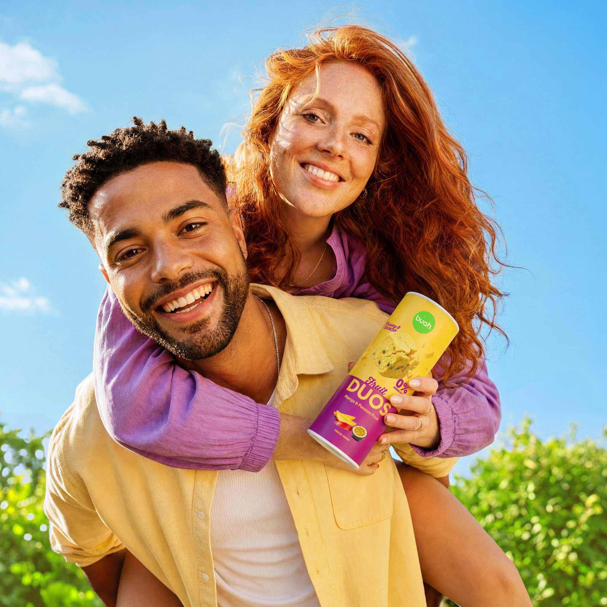 Ein fröhliches Paar an einem sonnigen Tag. Ein Mann trägt eine rothaarige, lächelnde Frau huckepack. Sie hält eine hohe Slim-Dose buah Fruit DUOS Mango & Passion Fruit in der Hand.