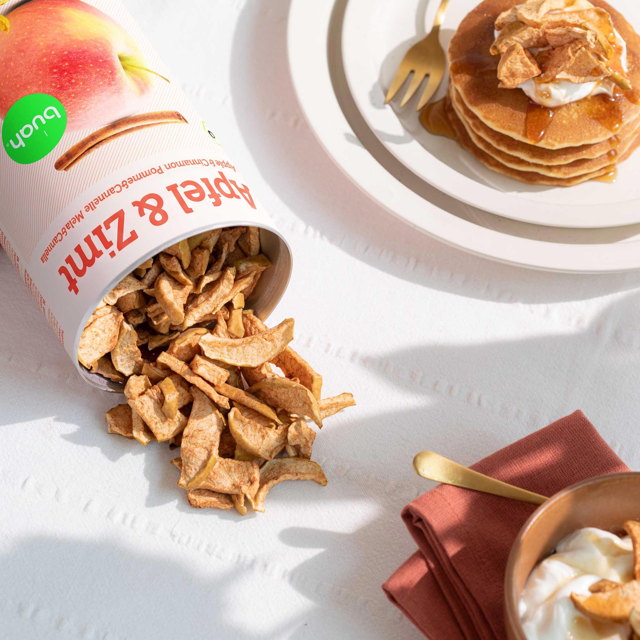 Eine auf dem Tisch liegende Dose buah gefriergetrocknete Früchte der Sorte Apfel & Zimt, aus der knusprige Apfelstücke auf eine weiße Tischdecke herausrieseln. Daneben stehen ein Teller mit garnierten Pancakes und eine Schale Joghurt im hellen Sonnenlicht.