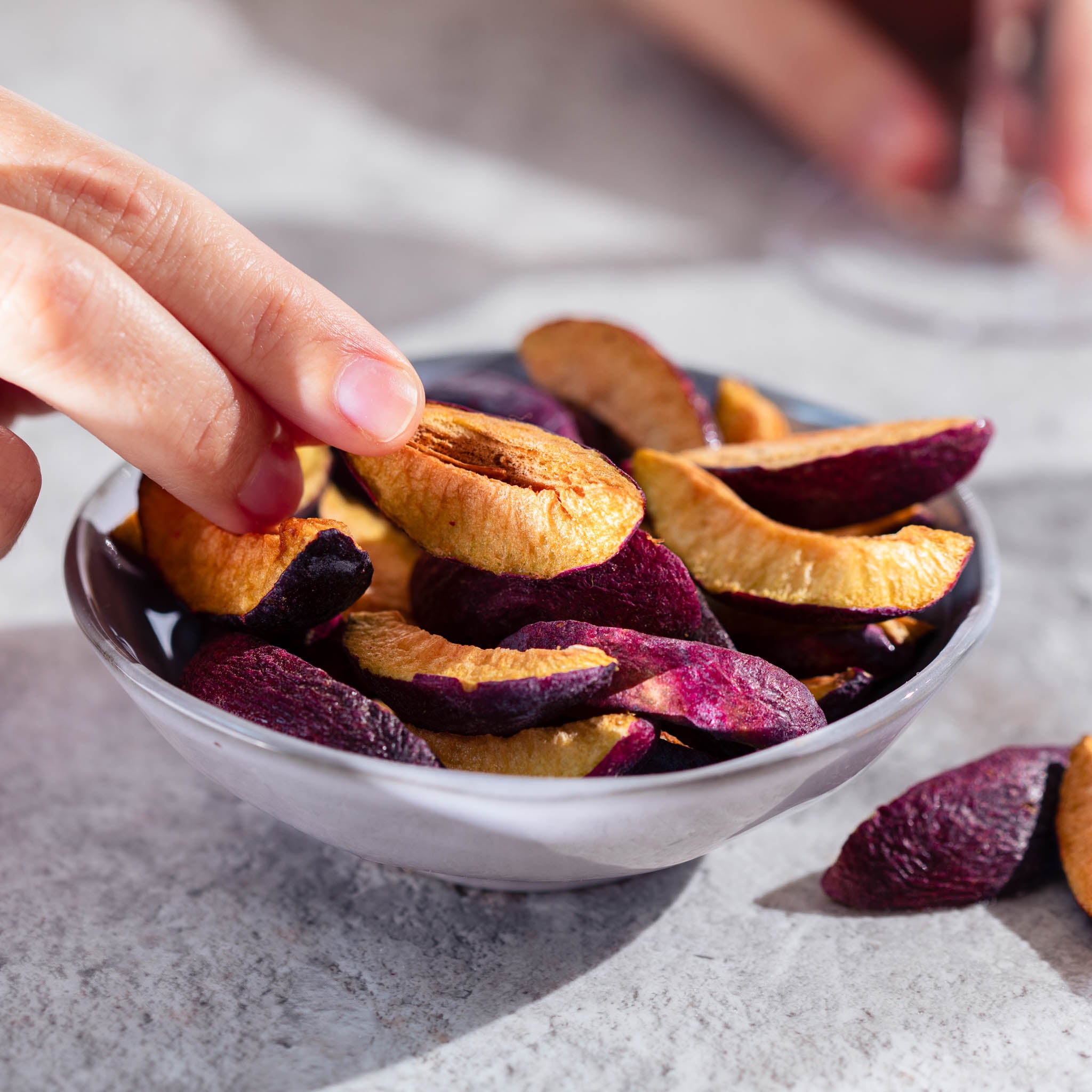 Hand greift in eine weisse Schale auf einem Tisch, voll mit buah gefriergetrockneter Pflaume in Orange und lila Farbe 