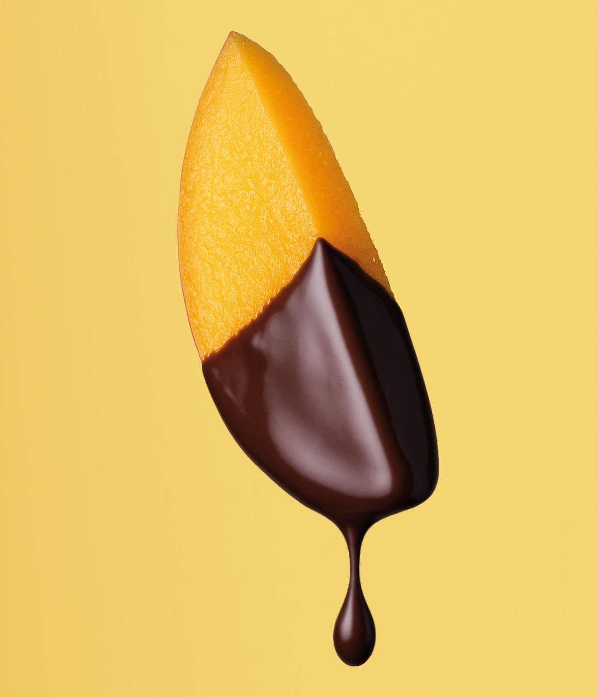 Eine gelbe Mangospalte wird in flüssige, dunkle 100% Kakaomasse getaucht vor einem sonnengelben Hintergrund